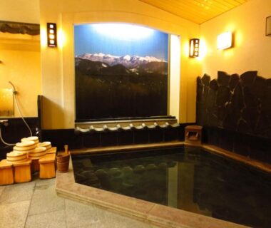 Indoor communal onsen with wooden stools, washing stations, stone-bordered bath and mountain mural at Shitanda in Gifu