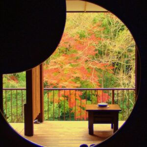 Circular window framing autumn maple trees, wooden veranda and low table at Shiunso ryokan in Kanagawa, Kantō