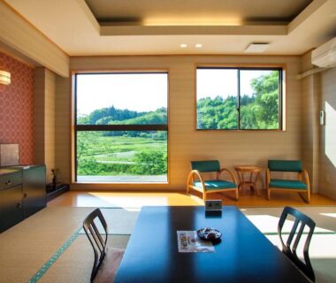 Bright tatami room with low dining table, chairs by large windows overlooking green fields and tree-covered hills