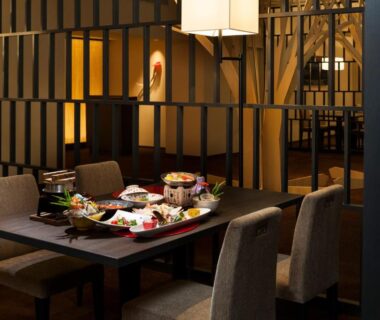 Indoor dining area with table set for Japanese dishes, partition screens, upholstered chairs, and warm ambient lighting