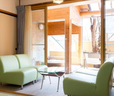 Light-filled tatami room with green sofas, low glass table, shoji panels and wooden bath beyond sliding doors