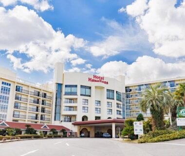 Facade of Hotel Hawaiians with palm trees, covered entrance and multi-story wings under blue sky in Fukushima