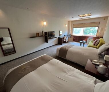 Bright twin room with two beds, seating area by large window and wooden media shelf at Suichokan Hokkaidō