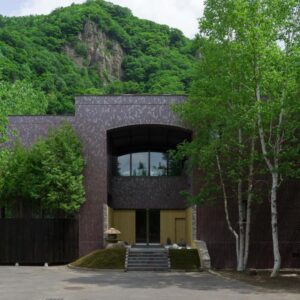 Brick façade of Suigan ryokan with arched window, stone steps and birch trees by forested hillside in Hokkaidō