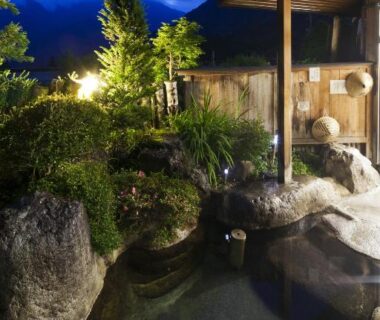 Rock-lined outdoor onsen bath with wooden roof, lantern-lit garden and mountain backdrop at Suisensou, Gifu