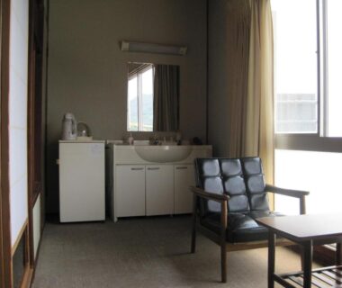 Compact ryokan room interior with washbasin, small refrigerator, seating area, shoji screen and bright window at Suminoe Ryokan, Hiroshima