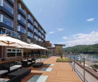 Outdoor wooden terrace with loungers and umbrellas beside a multi-story ryokan overlooking a coastal bay and hills