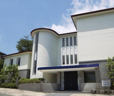 White two-story ryokan exterior with covered entrance, shrubs and driveway under clear blue sky in Hakone, Kanagawa