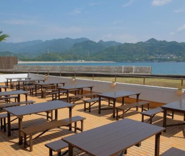 Outdoor wooden dining tables on seaside terrace with bay and distant mountains at Sunrise Katsuura Wakayama