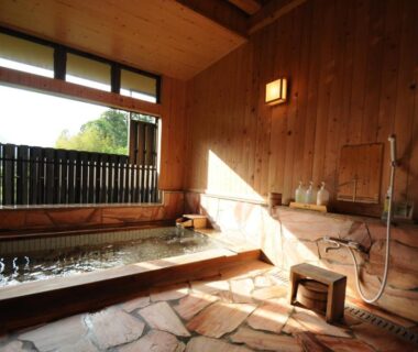 Indoor wooden bathing area with stone floor, soaking tub, hand shower and low stool overlooking hillside through large window
