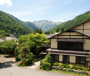 Traditional multi-story ryokan with exposed wooden beams, manicured greenery and mountains rising behind in Gunma Japan