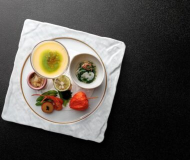 Ceramic bowl with clear broth and garnish, small bowls of greens and pickles on round tray over white cloth