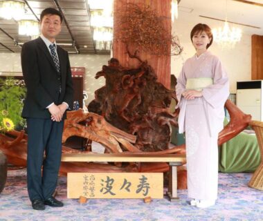 Woman in kimono and man in suit standing by wooden sculpture in ryokan lobby, Suzunami, Mie