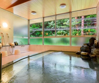 Indoor onsen with rock fountain, wooden ceiling, and large windows overlooking forest at Suzuranso, Nagano