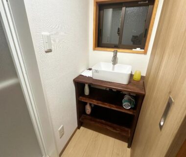 Compact washbasin on wooden vanity with open shelving, small window, light wood floor and striped bath mat