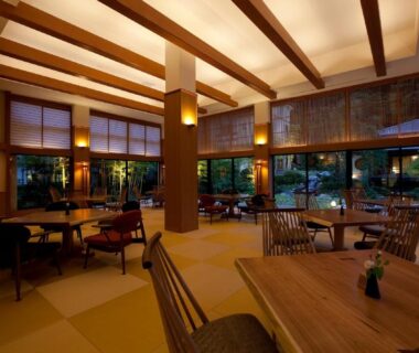Warm wooden lounge with tatami flooring, low tables and large windows with greenery at TAKITEI Riverside Onsen, Kanazawa