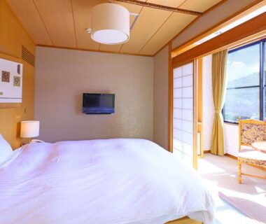 Sunlit room with low Western bed, tatami-style flooring, shoji doors and wooden seating by glass balcony