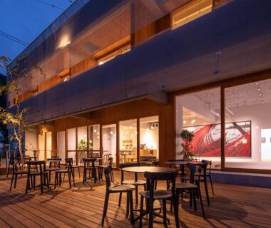 Wooden terrace with tables and chairs, warm-lit interior and artwork visible through large windows at TSUCHIYA Kinosaki onsen, Hyōgo