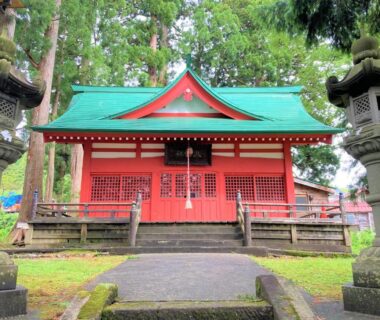 Small red Shinto shrine with green roof and stone lanterns beside wooded path at Tabataya, Niigata