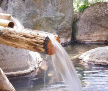 Wooden spout pouring water into stone-lined outdoor onsen pool with rocks and greenery at Tabist Kazeyuki, Gifu