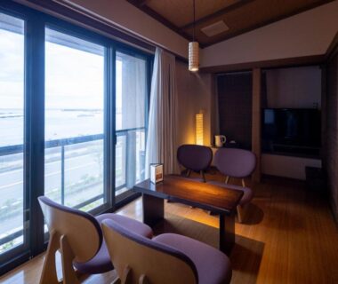 Ryokan sitting area with low table, wooden floors and sliding glass doors overlooking water and nearby road in Shizuoka