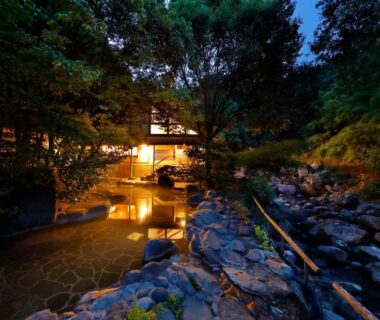 Stone outdoor onsen pool beside lit Taishoya building along river in Saga Kyushu