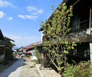 Traditional wooden ryokan facades lining a stone-paved lane with greenery under a clear blue sky in Gifu, Chūbu