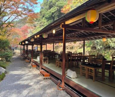 Covered wooden riverside dining terrace with lanterns, tables and autumn foliage at Takao Kanko Hotel Kyoto