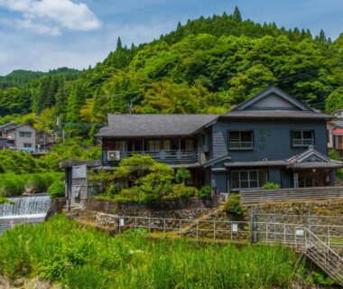 Two-story wooden ryokan beside river and stepped weir with lush green hillside backdrop in Ōita