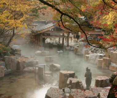 Rock-lined outdoor onsen pools with steam, maple foliage and wooden bridge at Takaragawa Onsen Ousenkaku Gunma