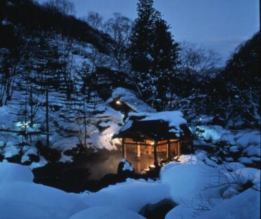Lit wooden onsen pavilion at Takaragawa Onsen Ousenkaku by snowy riverside at dusk in Gunma