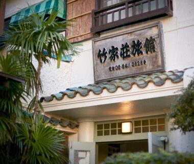 Wooden sign and tiled eave above the Takeyaso Ryokan entrance framed by greenery in Hyōgo, Kansai