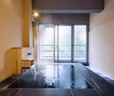 Stone-tiled indoor onsen bath with wooden stool and flowing faucet at Tamana Onsen Happouen, Kumamoto, Kyūshū, Japan