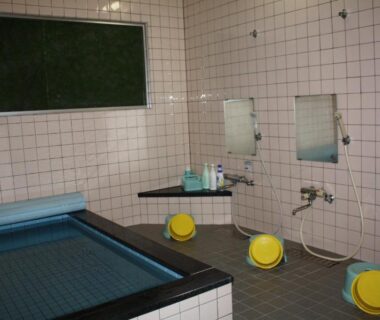 Tiled communal bathing room with washing stations, yellow stools, mirrors and covered soaking tub at Tamonkan, Yamagata