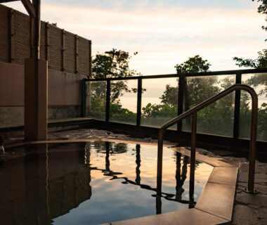 Stone-lined outdoor onsen with handrail and glass barrier overlooking trees and distant hills