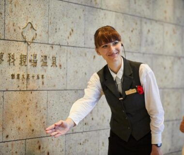 Smiling receptionist in uniform guiding a guest in the ryokan lobby by a stone wall sign