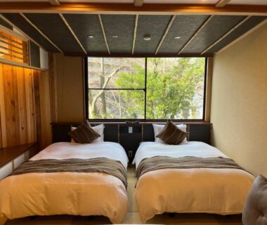 Twin low beds on tatami floor with wooden accents and large window overlooking garden trees at Tatsumikan, Gunma