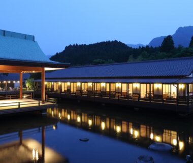 Evening view of illuminated wooden ryokan buildings reflected in calm pond at Tawarayama Onsen Ryokan Minawa Kumamoto