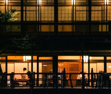 Warmly lit wooden facade with guests dining on balcony behind shoji screens at Tazuru ryokan in Kyoto