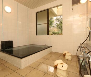 Tiled indoor bathing room with rectangular tub, wooden stools and buckets, shower fixtures and window with outside greenery