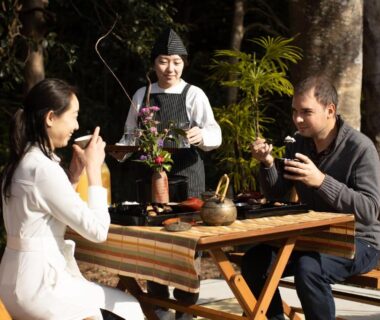 Guests enjoying tea at a wooden outdoor table with a server and trees at Temple Hotel 正暦寺 Kyoto