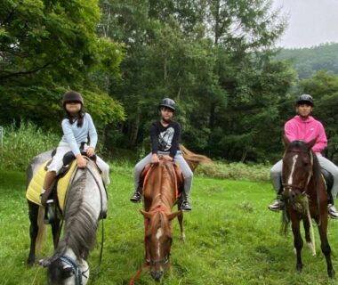 Three riders in helmets on horses in a grassy clearing with forested hills at a Nagano ryokan