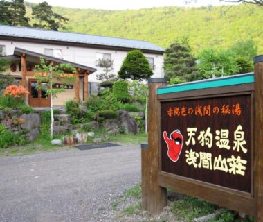 Wooden signboard with ryokan name in Japanese, landscaped entrance, gravel driveway and building set against green hills