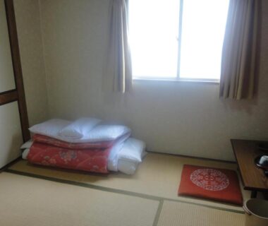 Stacked futon on tatami floor beside low table and curtained window at Tenryu Ryokan Hiroshima