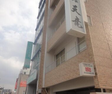Multi-story brick ryokan exterior with vertical Tenryu Ryokan sign, street entrance and parked car in urban Hiroshima neighborhood