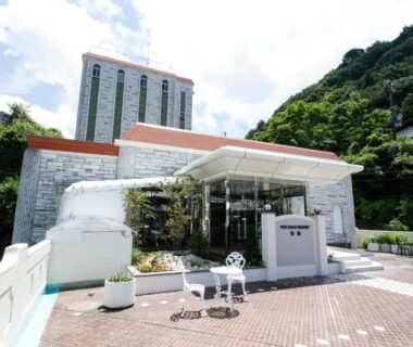 Glass entrance and terrace seating at The Gran Resort Arima with hillside hotel tower and paved courtyard in Hyōgo