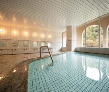 Tiled indoor onsen pool with arched windows overlooking wooded hillside at The Gran Resort Kinosaki in Hyōgo