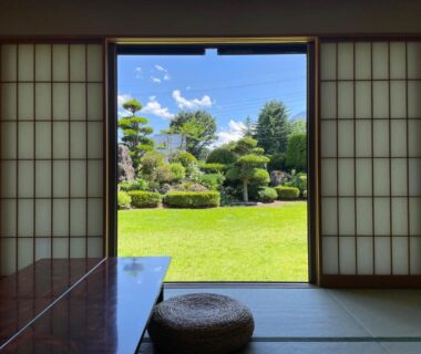 Tatami room with shoji screens, low table and round cushion opening onto a trimmed garden under blue sky in Yamanashi