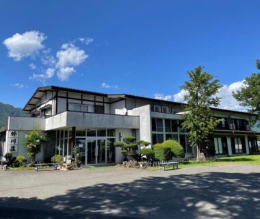 Two-story ryokan building with manicured shrubs, mountain backdrop and clear blue sky at Togawaso in Yamanashi