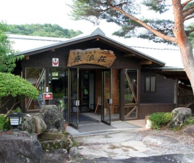 Entrance of Tokonamiso ryokan with wooden facade, stone path, pine tree and shrubs in Nagano